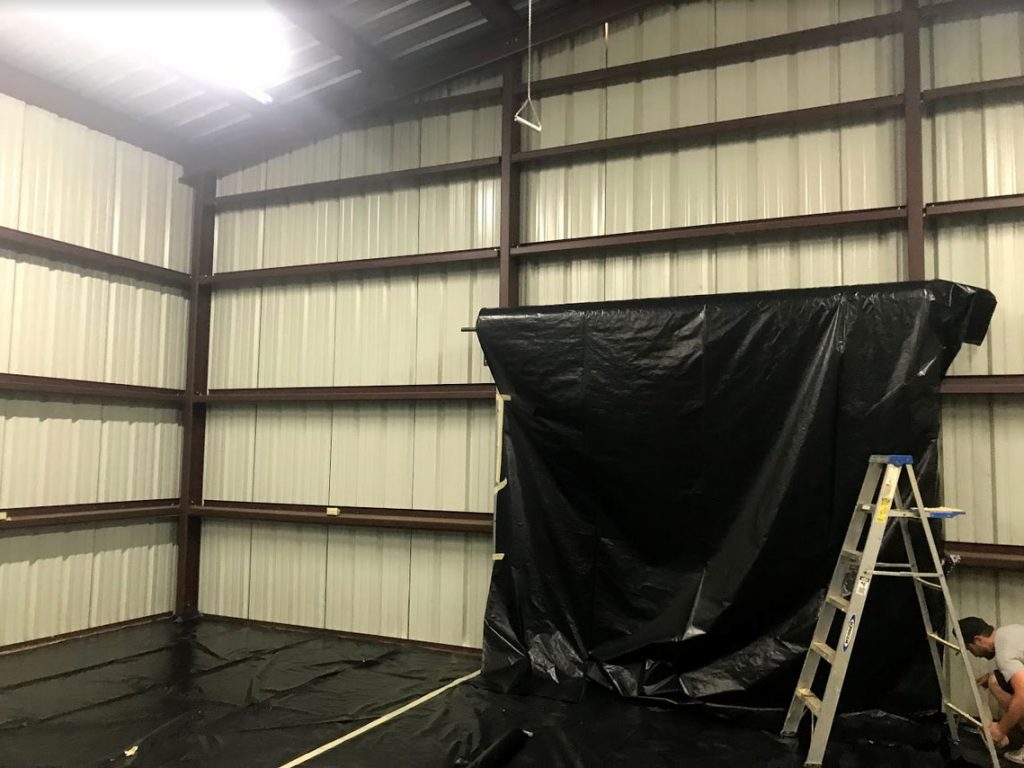 The interior of a metal-framed room features a ladder and black plastic sheets partially covering the walls and floors, hinting at the recent work by an insulation installer. This space has been prepped for high-quality spray foam insulation.